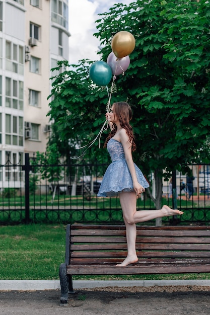 A ballerina in a corset dress walks on tiptoe on a bench in the park holding balloons in her hands