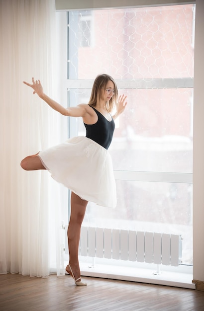 Ballerina girl in glasses stands on one leg near the window in the afternoon in a bright studio