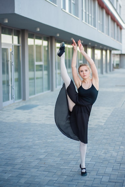 Балерина в пачке позирует на фоне жилого дома Красивая молодая женщина в черном платье и пуантах прыгает, демонстрируя растяжку Балерина стоит в шпагате Снаружи