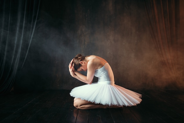 ballerina in white dress sitting on theatrical stage, side view. classical ballet dancer training in class on black