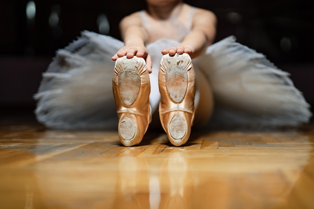 Ballerina zittend op de vloer van de klas en en bedrijf voor tiptoes van witte balletschoenen op de houten vloer. klassieke ballerina's 'pointes van een klein meisje. detailopname