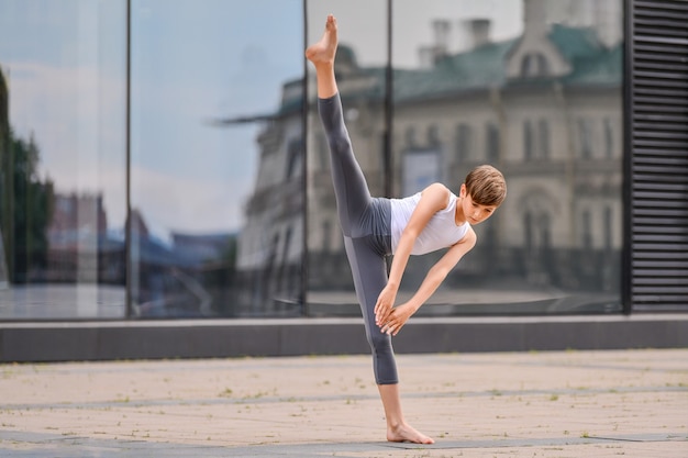 L'adolescente del ragazzo di balletto si allena in piedi sullo sfondo del riflesso della città e del cielo nella parete di vetro