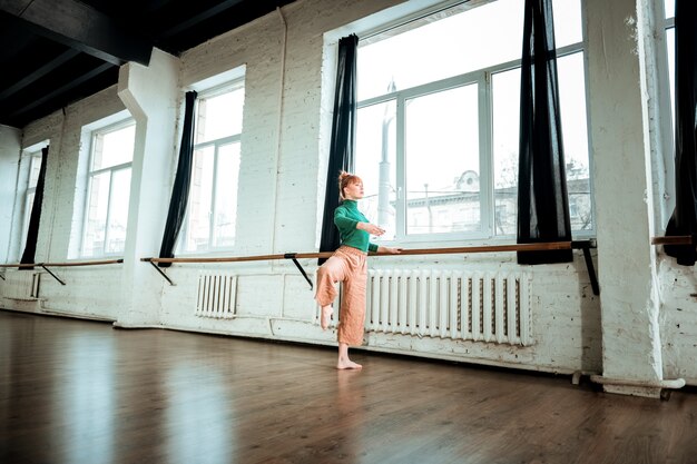 Foto scuola di balletto. giovane ballerino moderno professionista con i capelli rossi che sembra serio durante l'allenamento vicino al bar di balletto