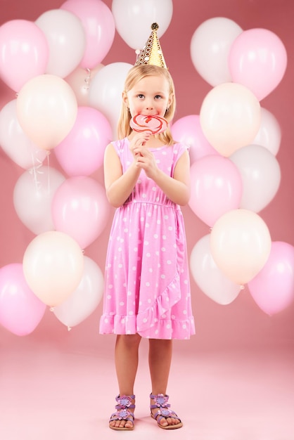 생일 파티 축하 및 특별한 날을 위해 스튜디오에 있는 소녀의 초상화와 릴리팝 풍선, 핑크색 배경에 사탕과 달한 과자 및 디저트와 함께 행복하고 흥분하고 사랑스러운 어린 아이의 미소