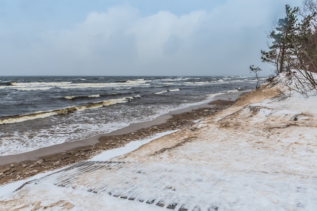 Пляж Балтийского моря зимой снежный, а в море большие волны. Пешеходная дорожка между зимними дюнами Балтийского моря в Саулкрасты в Латвии