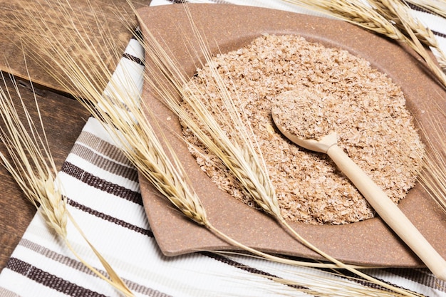 Бамбуковая тарелка и деревянная ложка, наполненная пшеничными отрубями, в окружении колосьев пшеницы