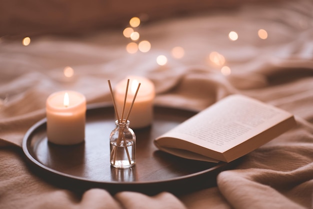 Bamboo sticks in bottle with scented candlrs and open book on wooden tray in bed closeup. Home aroma.