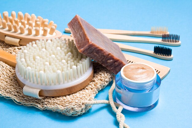 Bamboo toothbrushes, wooden comb, knitted washcloth, natural homemade scrub in a jar and homemade soap and dry massage brush on a blue surface