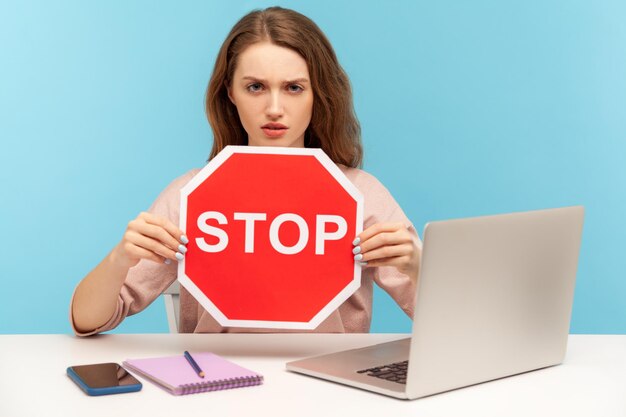 Ban, forbidden access. Young woman holding stop symbol, warning with red traffic sign and looking angrily, sitting at workplace with laptop, home office. indoor studio shot isolated on blue background