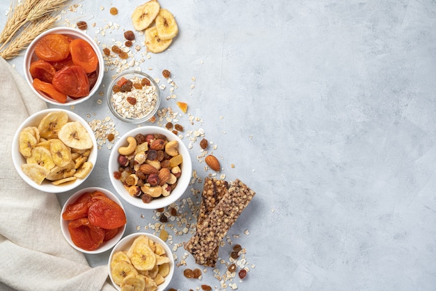 Banana chips, dried apricots, different types of nuts, raisins and granola bar inside white bowls