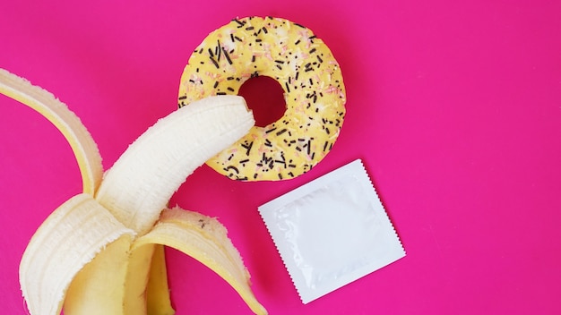Банан, пончик и презерватив. Идея секса. Яркая картинка на розовом фоне. Понятие здоровья и защищенного секса