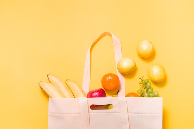 Bananas, tangerines, grapes, apple, pear and lemons in a fabric shopping bag on yellow surface