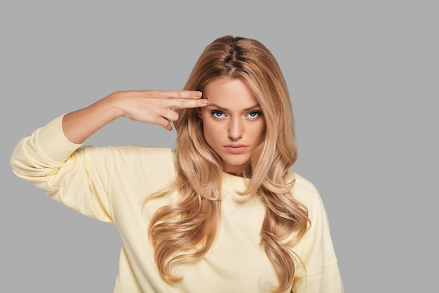 Bang! Attractive young woman gesturing handgun near head and looking at camera while standing against grey background