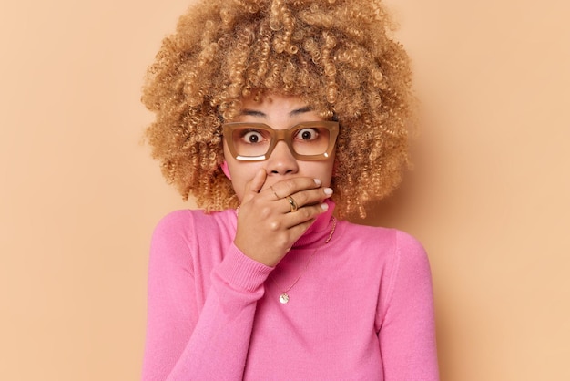 Foto bang bezorgde jonge vrouw hapt naar adem van angst bedekt mond met hand ziet verschrikkelijk ongeval gekleed in casual roze coltrui draagt een bril geïsoleerd over roze achtergrond houdt adem in van schrik