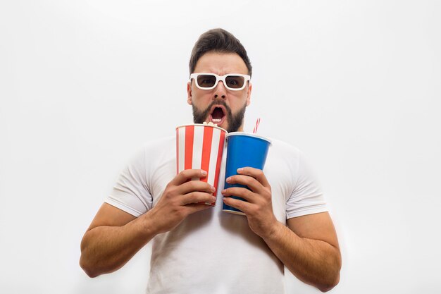 Bang en zorgen jongeman staat en houdt emmer popcorn met glas cola. Hij is erg bang. Jonge man houdt mond open.