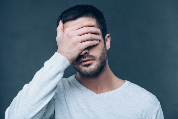 バン！欲求不満の若い男が頭の近くで鉄砲のジェスチャーを身振りで示し、灰色の背景に立っている間目を閉じたまま
