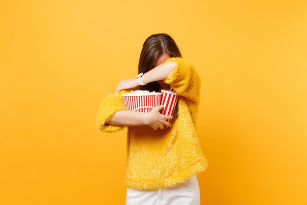 Bang jonge vrouw met lowere hoofd in 3d imax bril kijken naar film film verbergen die betrekking hebben op gezicht met arm houden popcorn kopje frisdrank geïsoleerd op gele achtergrond. Mensen oprechte emoties in de bioscoop.