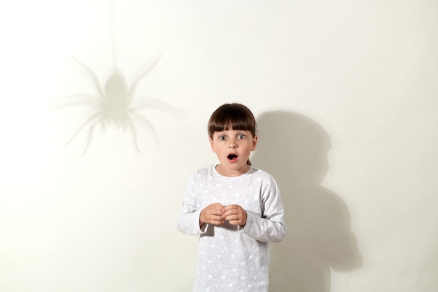 Bang meisje met een wit casual shirt dat weer poseert op de grijze muur met schaduw van spin, vrouwelijk kind met wijd geopende mond, bewegingloos staand, fobie voor insecten.