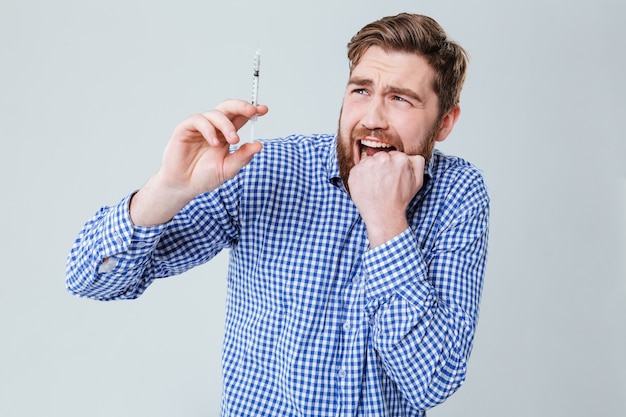 Bange, bebaarde jonge man die een spuit vasthoudt en in de zijne bijt