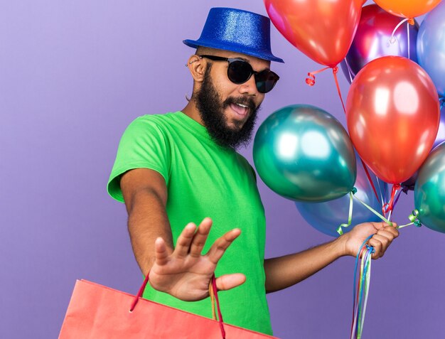 Bange jonge afro-amerikaanse man met een feestmuts met een bril die ballonnen vasthoudt met een cadeauzakje met stopgebaar geïsoleerd op een blauwe muur