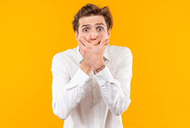 Bange jonge knappe kerel met een wit overhemd bedekte mond met handen geïsoleerd op een oranje muur