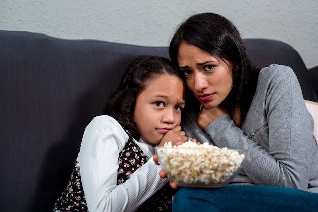 Bange meisjeszusters die op de bank zitten die de camera bekijken. Tv-horrorfilm kijken samen met popcorn.