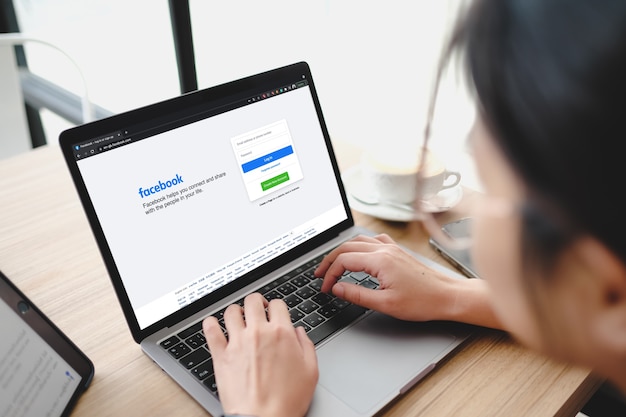 バンコク。タイ。 6月1,2021-カフェでビジネスパーソンの手にあるラップトップコンピューターデバイスのアプリ画面にログイン、サインアップ登録ページにfacebookソーシャルメディアアプリのロゴ。