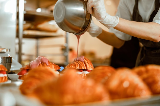 Banketbakker giet roze glazuur op smakelijke croissant uit metalen kom op aanrecht