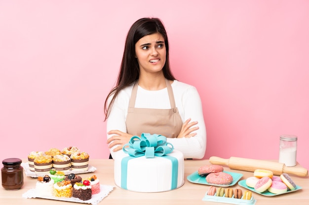 Banketbakker met een grote cake in een tafel over geïsoleerde roze muur met verwarde gezichtsuitdrukking