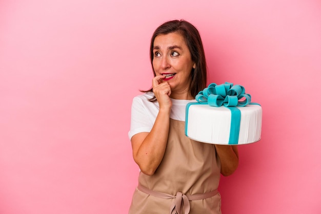 Banketbakker vrouw van middelbare leeftijd met een taart geïsoleerd op blauwe achtergrond ontspannen denken over iets kijken naar een kopie ruimte.