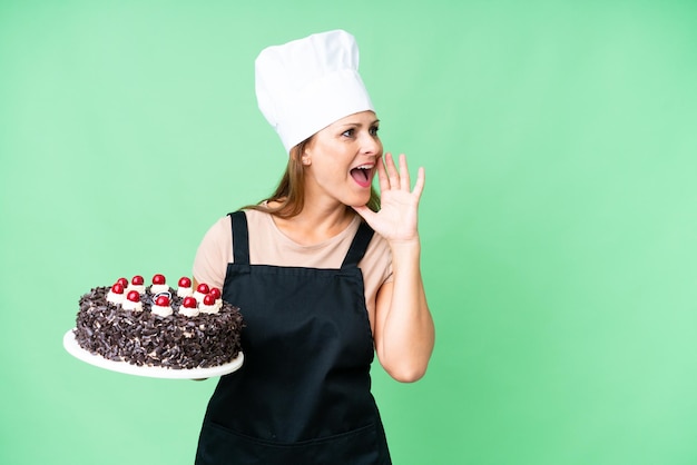 Banketbakkersvrouw van middelbare leeftijd die een grote cake vasthoudt over geïsoleerde achtergrond schreeuwend met wijd open mond naar de zijkant