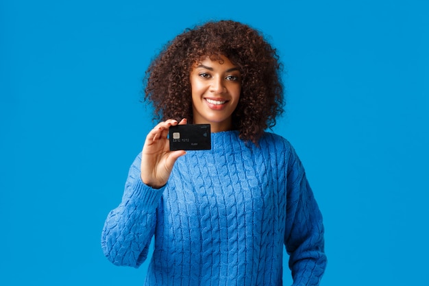 Concetto bancario, commerciale e finanziario. attraente donna afro-americana piacevole con capelli afro, maglione invernale, mostrando la carta di credito, pagando l'acquisto online, prepararsi per le vacanze