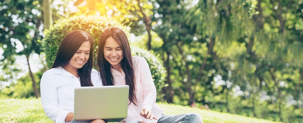 Photo banner asian two women talking using laptop sit on green meadow at green park panorama friendship girls chat talk look at laptop small business beautiful women laugh talk together with copy space