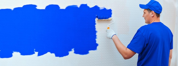 배너 건설 및 수리 제복을 입은 남자가 페인트 롤러로 방 안의 벽을 파란색으로 칠합니다