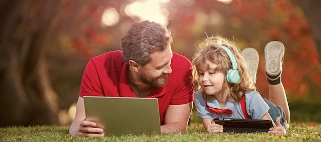 Banner of father on son school boy with laptop study online\
lying on grass dad and son use laptop for video call or lesson\
listen music in headphones in park education online