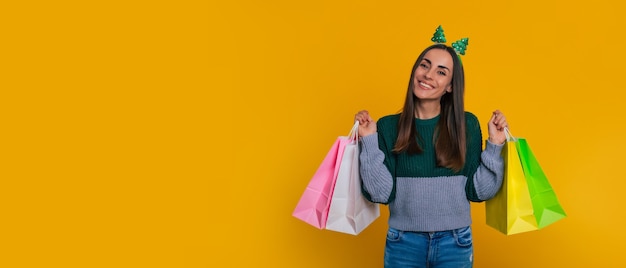 Banner foto van gelukkige aantrekkelijke jonge brunette vrouw in kerstinkopen tijd met kleurrijke papieren zakken voor aankoop en geschenken geïsoleerd op gele achtergrond