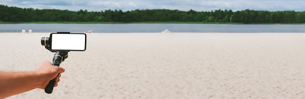 Banner, Mockup van een smartphone op een Steadicam in de hand van een man. Tegen de achtergrond van een zandstrand en de natuur met een meer.