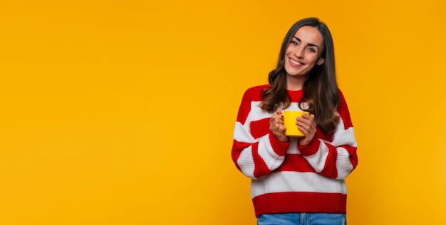 黄色の背景に分離された手で熱いお茶やコーヒーの黄色のカップとかわいい幸せな若いブルネットの女性のバナー写真