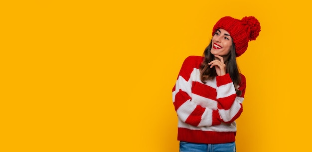 Фотография баннера симпатичной современной счастливой брюнетки в красной зимней шапке и шарфе и позирующей изолированной на желтом фоне