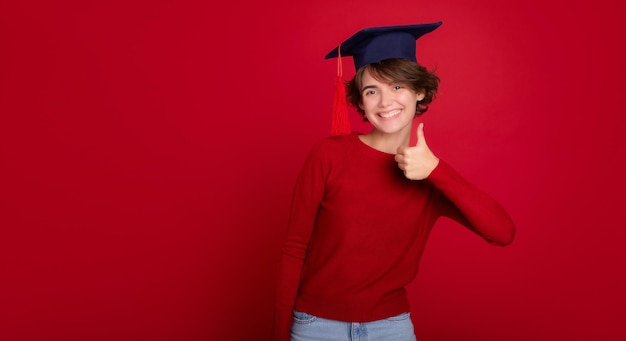 Banner photo of happy pretty excited woman graduate with masters cap shows her thumb up isolated in studio on red background University study education college knowledge graduation