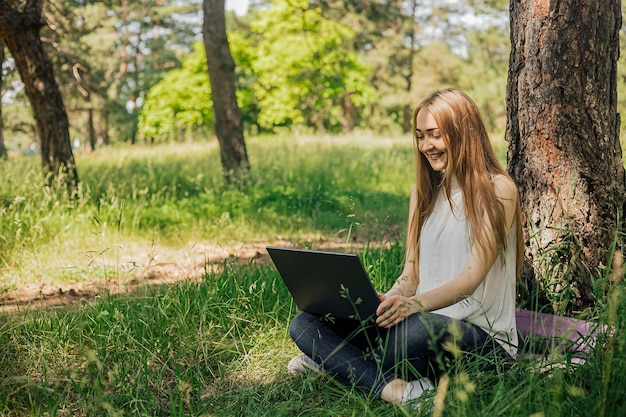 배너에서 어린 소녀는 잔디밭에 앉아 있는 공원의 신선한 공기 속에서 노트북을 가지고 일합니다. 원격 작업의 개념 프리랜서로 일합니다. 소녀는 노트북으로 강의를 듣고 미소를 짓습니다