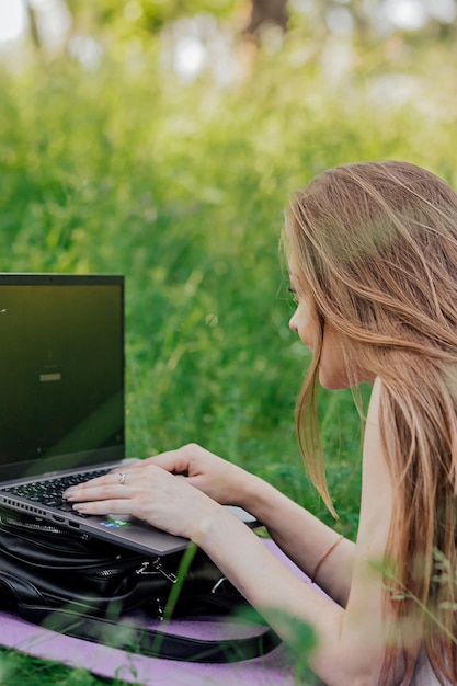 배너에서 어린 소녀는 잔디밭에 앉아 있는 공원의 신선한 공기 속에서 노트북을 가지고 일합니다. 원격 작업의 개념 프리랜서로 일합니다. 소녀는 노트북으로 강의를 듣고 미소를 짓습니다