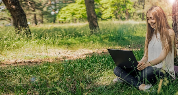 Photo on the banner a young girl works with a laptop in the fresh air in the park sitting on the lawn the concept of remote work work as a freelancer the girl takes courses on a laptop and smiles