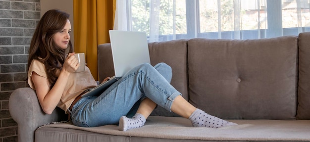 A banner of a young woman is drinking a cup of coffee during working from home A girl is thinking and sitting on the sofa with a laptop