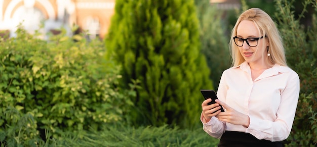 Bannerportret van een mooie vrolijke zakenvrouw in een shirt en zwarte rok die haar smartphone gebruikt, zit tegen de achtergrond van een bloembed