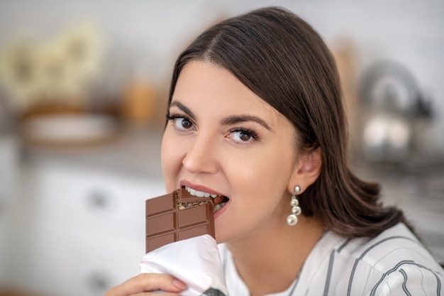 チョコレートバー。チョコレートを食べる縞模様のブラウスの黒髪の女性