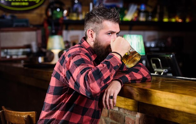 Bar is relaxing place have drink and relax Man with beard spend leisure in dark bar Hipster relaxing at bar with beer Brutal hipster bearded man sit at bar counter drink beer Order alcohol drink