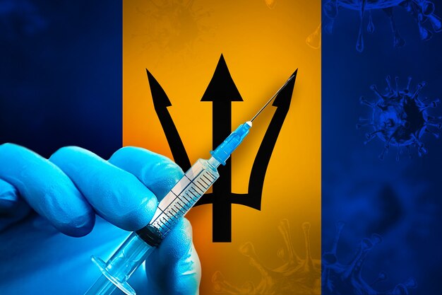 Кампания по вакцинации против Covid19 на Барбадосе Рука в синей резиновой перчатке держит шприц перед флагом