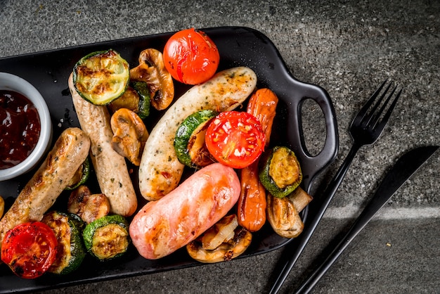 Barbecue. Assortiment van verschillende gegrilde vleesworsten, met groenten-BBQ - champignons, tomaten, courgette, uien. Op een zwarte stenen tafel, op een zwarte plaat, met saus. Kopieer ruimte bovenaanzicht