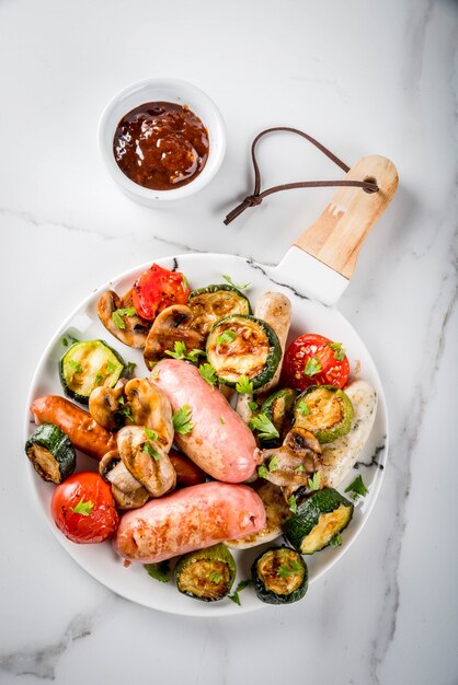 Barbecue. Assortment of various grilled meat sausages, with vegetables BBQ  mushrooms, tomatoes, zucchini, onions. On white marble table, on plate, with sauce. copyspace top view